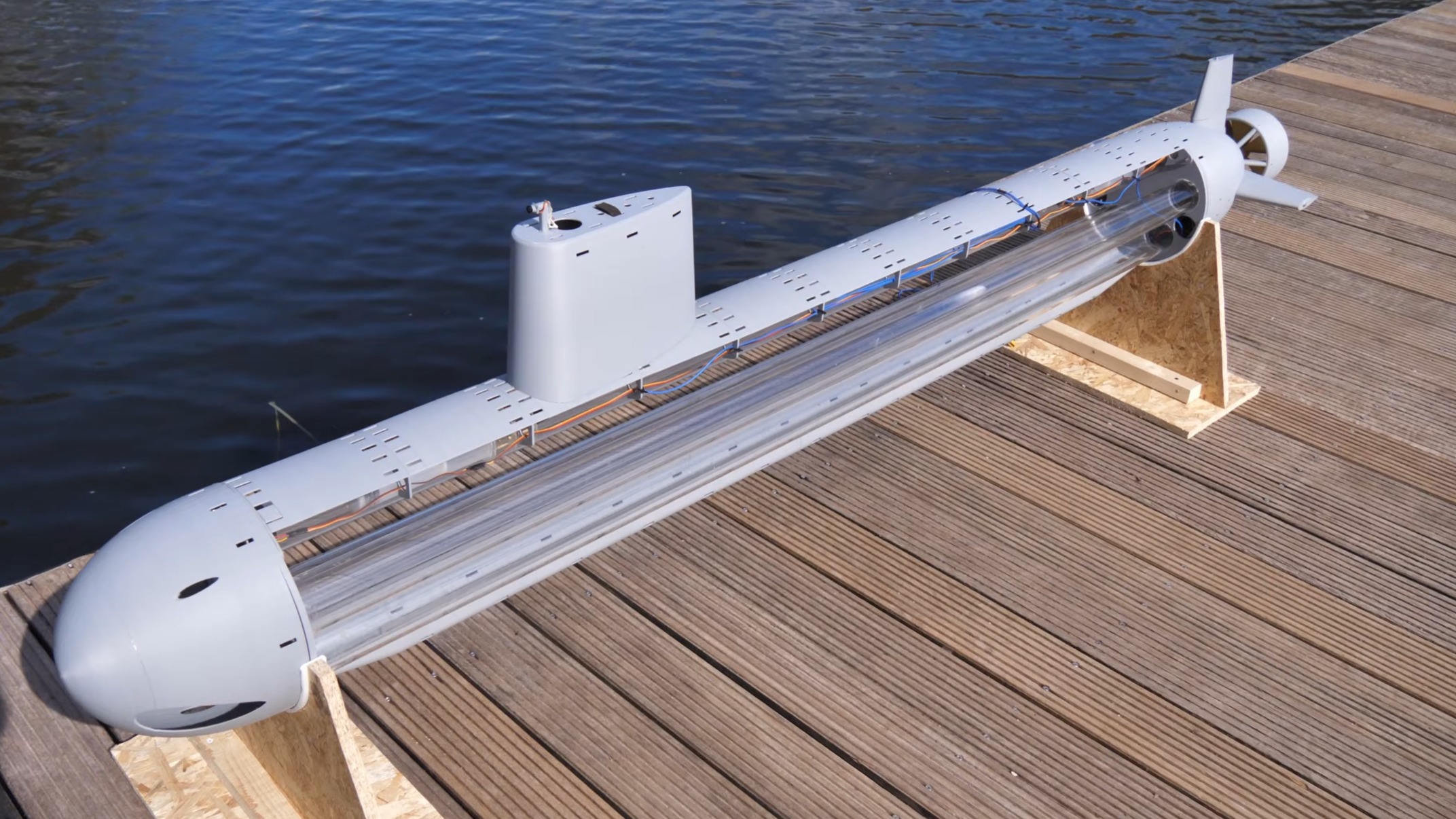 A model submarine is shown on a dock. The body of the submarine is made out of a series of acrylic tubes, with other parts made out of grey plastic.