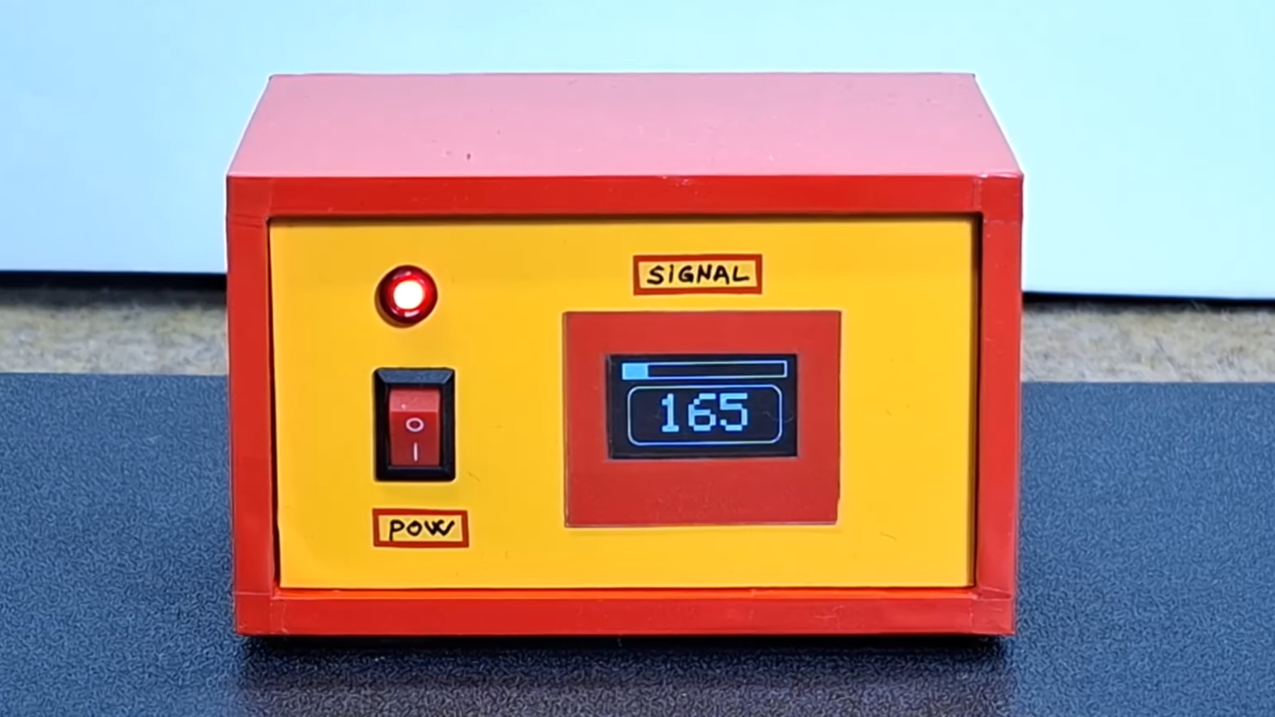 A red box with a yellow front panel is shown. The front panel contains a power switch, an indicator light, and a small OLED display.